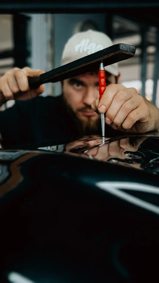 Dent-repair technician working on a glossy vehicle panel with precision tapping tools — example stock photo, not BumperFixers work