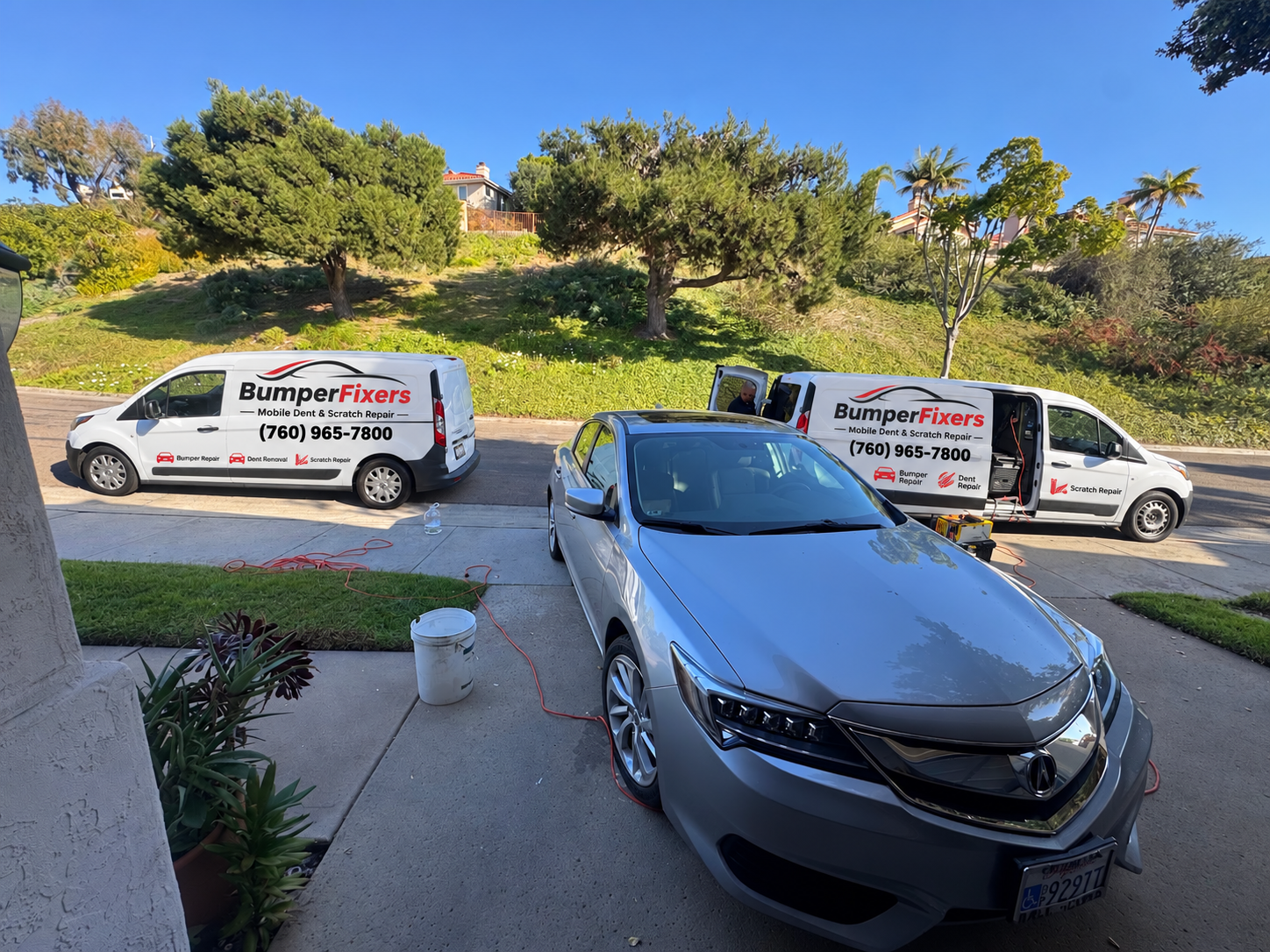 Two Bumper Fixers vans working on a silver Acura in a customer driveway