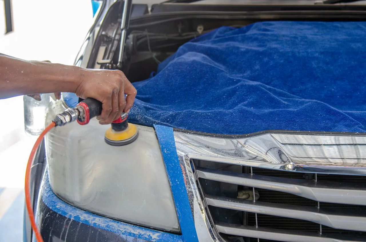 Worker machine-polishing a cloudy headlight with blue masking tape protecting surrounding paint — example stock photo, not BumperFixers work